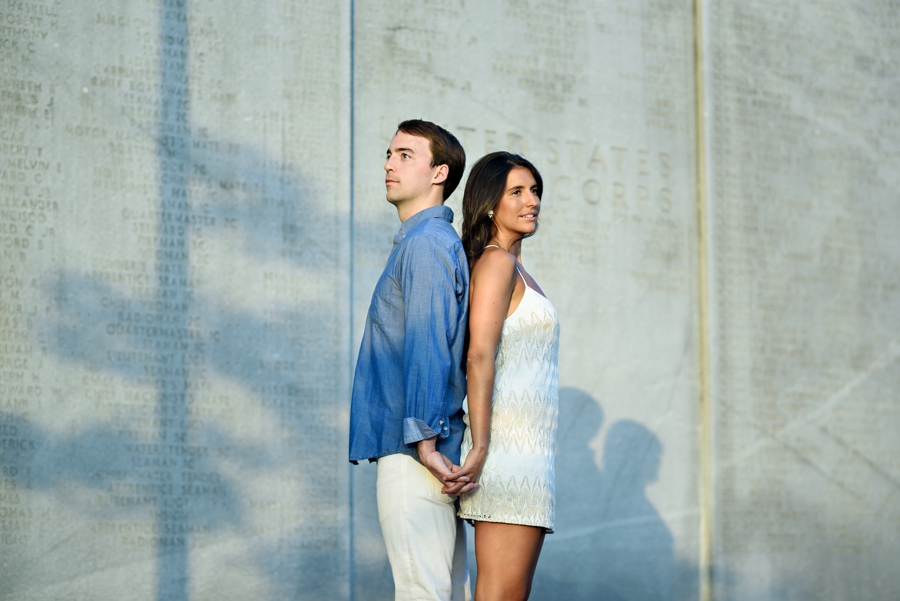 A sexy couple stand back to back and hold hands in the sun during their engagement session in battery park nyc.